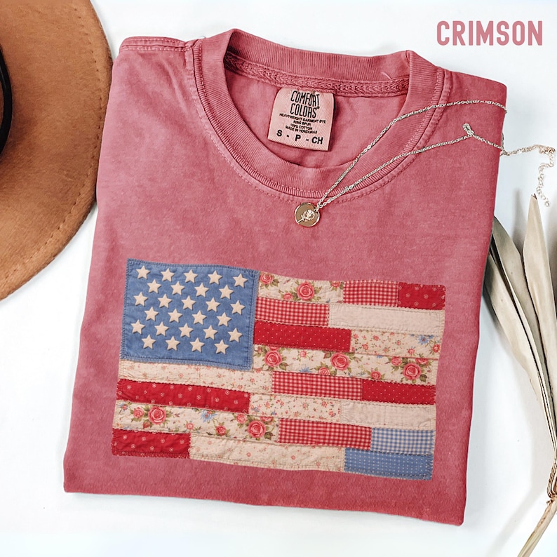 May include: Crimson-colored t-shirt with a patchwork American flag design. The flag features a blue field with white stars and red and floral-patterned stripes. A silver necklace with a pendant is draped on the shirt.