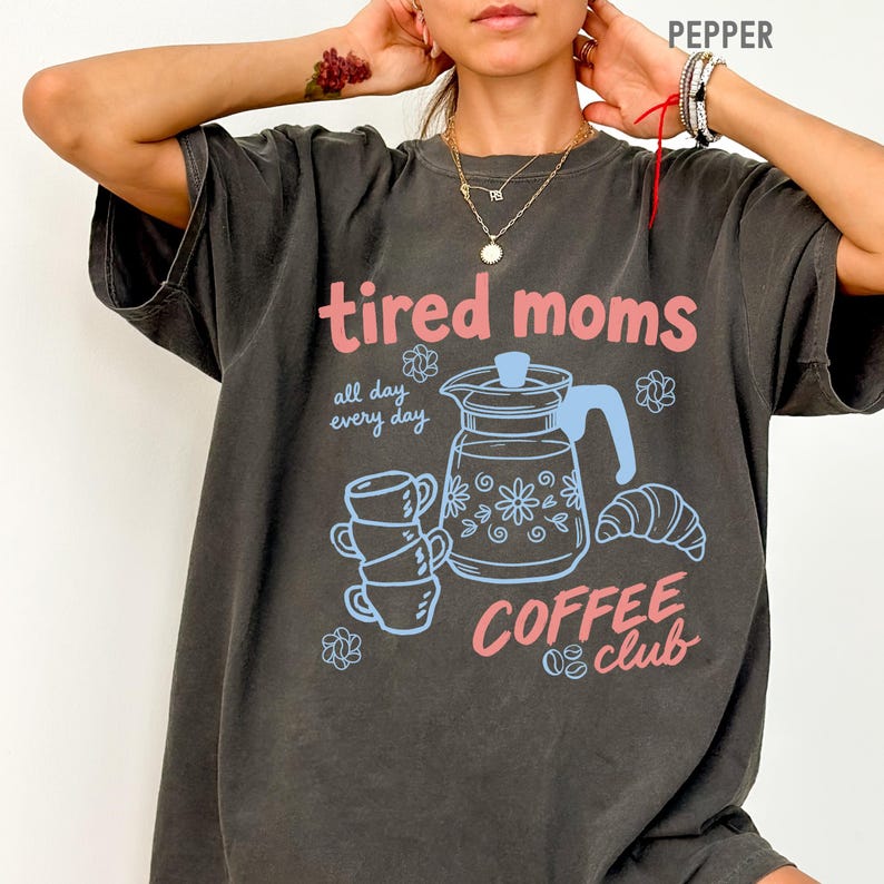 Vintage style graphic t-shirt featuring coffee pot, stacked cups, croissant, and playful tired moms coffee club text in soft pastel colors for moms.