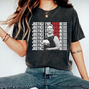 May include: Dark gray t-shirt with the text "JUSTICE FOR GOODNESS" in white and red. A black and white photo of a woman holding flowers is on the shirt. The shirt is worn with blue jeans.