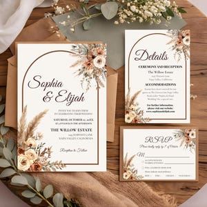 May include: A wedding invitation suite featuring a main invitation, a details card, and an RSVP card. Each piece has a white background, a brown arched border, and floral accents with pampas grass, dried flowers, and roses in shades of cream and brown.