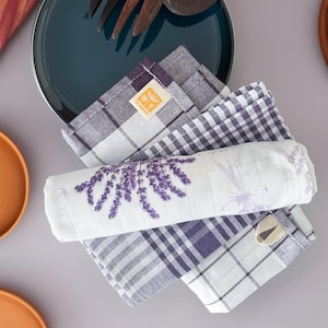 May include: A stack of purple and white checkered and floral patterned kitchen towels. One towel is rolled up, displaying a lavender design. The towels are arranged near a dark blue plate and wooden utensils.