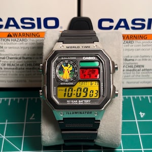 Könnte beinhalten: Eine digitale Casio-Uhr mit schwarzem Armband und silberfarbenem Gehäuse. Das Zifferblatt zeigt die Uhrzeit, das Datum und eine Pikachu-Grafik an. Die Uhr trägt die Aufschrift "WR100M" und "10 YEAR BATTERY". Die Uhr liegt auf einer weißen Oberfläche.