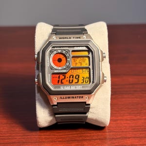 May include: A digital Casio watch with a black strap and silver-tone case. The watch face shows the time, date, and world time features. The watch has the text "WORLD TIME", "CASIO", "WR100M", "10 YEAR BATTERY", and "ILLUMINATOR".