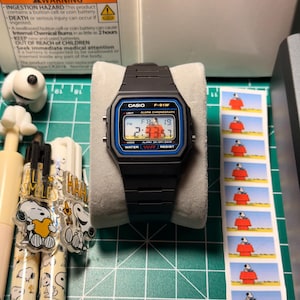 May include: A black Casio F-91W digital watch with a blue and red display, on a white cushion. The watch shows the time and date. The image also includes Casio packaging, Snoopy-themed pens, and Snoopy stamps.