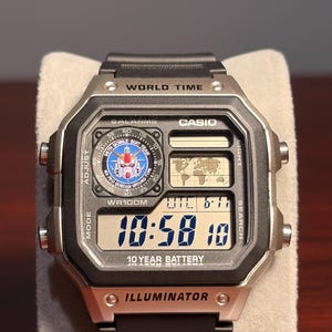 Puede incluir: Un reloj digital Casio con caja negra y plateada y correa negra. La esfera del reloj muestra la hora, la fecha y las funciones de hora mundial. El reloj tiene el texto "10 YEAR BATTERY" e "ILLUMINATOR".