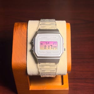 Op de afbeelding: Een Casio F-91W digitale horloge met een transparante band en zilveren kast. De witte wijzerplaat toont de tijd, datum en dag in roze en paars. Het horloge wordt gepresenteerd op een houten horlogestandaard.