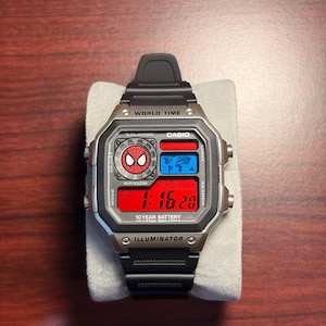 May include: A black and silver Casio digital watch with a red digital display showing the time. The watch features a Spider-Man logo and the text "WORLD TIME" and "ILLUMINATOR". The watch has a black band and is displayed on a white surface.