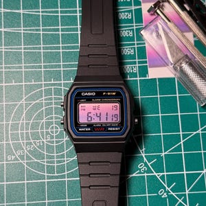 Op de afbeelding: Zwarte Casio F-91W digitale horloge met een zwarte band. De wijzerplaat toont de tijd, dag en datum. Het horloge ligt op een groene snijmat. De wijzerplaat heeft de tekst "CASIO F-91W" en "WATER RESIST".