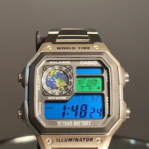 May include: A silver and black Casio digital watch with a blue digital display showing the time, date, and world map. The watch features the text "WORLD TIME", "5 ALARMS", "WR100M", and "10 YEAR BATTERY".