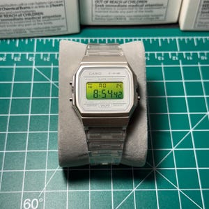 Könnte beinhalten: Eine silberne Casio F-91W Digitaluhr mit einem transparenten Kunststoffarmband. Das Zifferblatt zeigt die Uhrzeit in grünen Ziffern an. Die Uhr liegt auf einer grauen Oberfläche vor einem grünen Gitterhintergrund.