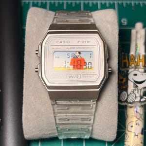 May include: A Casio F-91W digital watch with a transparent plastic strap and a silver-coloured case. The watch face shows the time and a cartoon graphic. The watch is on a grey surface.