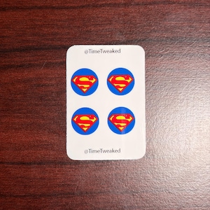 May include: A white rectangular sticker featuring four blue circles with the Superman logo in the center. The Superman logo is a red and yellow "S" inside a yellow diamond. The text "@TimeTweaked" is printed at the top and bottom of the sticker.