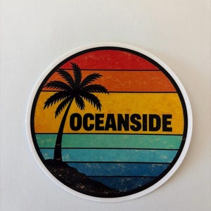 May include: A circular sticker featuring a retro-style sunset with horizontal bands of red, orange, yellow, teal, and blue. A black silhouette of a palm tree stands on a small hill on the left side, with the word OCEANSIDE printed in black text.