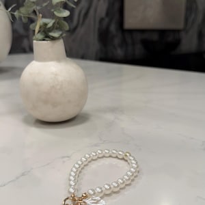 May include: A circular bracelet composed of small white spherical beads. The bracelet features a gold-toned metal clasp and a collection of charms, including a single larger white pearl bead and a textured, irregular white disc-shaped charm.