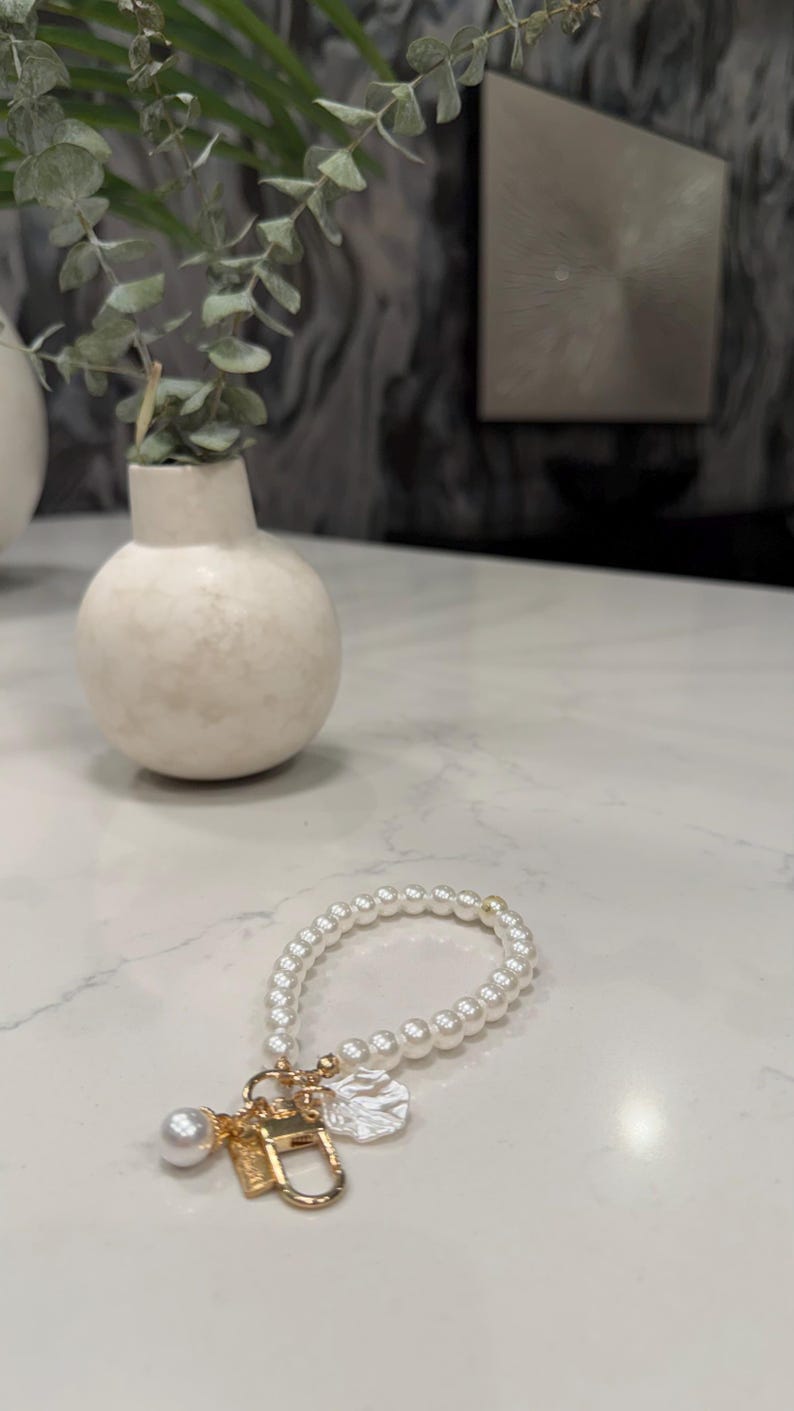 May include: A circular bracelet composed of small white spherical beads. The bracelet features a gold-toned metal clasp and a collection of charms, including a single larger white pearl bead and a textured, irregular white disc-shaped charm.