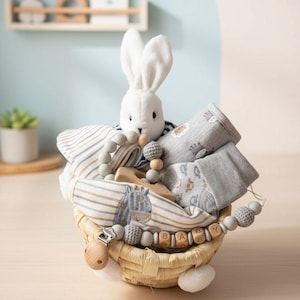 May include: A baby gift basket with a white stuffed bunny, baby clothes, wooden toys, and a pacifier clip. The basket is made of woven material and contains gray and white striped blankets and socks. The pacifier clip spells out "BABY".