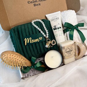 May include: A gift box with a variety of items, including a green velvet pouch with "Mom" in gold lettering, a pearl bracelet, hand cream, a candle, a hairbrush, and a white towel tied with a green ribbon. The box is brown and has the text "Calm Nest Box".