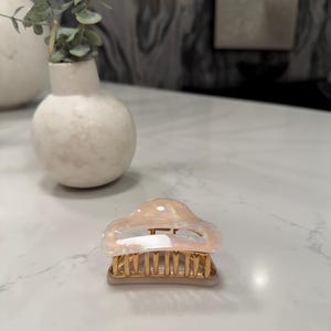 May include: A hair claw clip featuring a translucent, iridescent, pearlescent top section with a smooth, rounded shape. The base of the clip is constructed from gold-toned metal with a series of vertical teeth designed for securing hair in place.