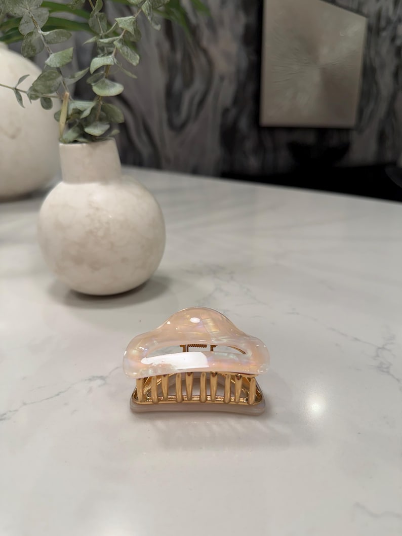 May include: A hair claw clip featuring a translucent, iridescent, pearlescent top section with a smooth, rounded shape. The base of the clip is constructed from gold-toned metal with a series of vertical teeth designed for securing hair in place.