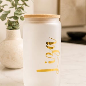 May include: A cylindrical frosted glass tumbler with a light-colored bamboo lid. The glass features the name Liza written in a metallic gold cursive script font on the front surface. The tumbler has a smooth, matte finish and a straight-sided design.
