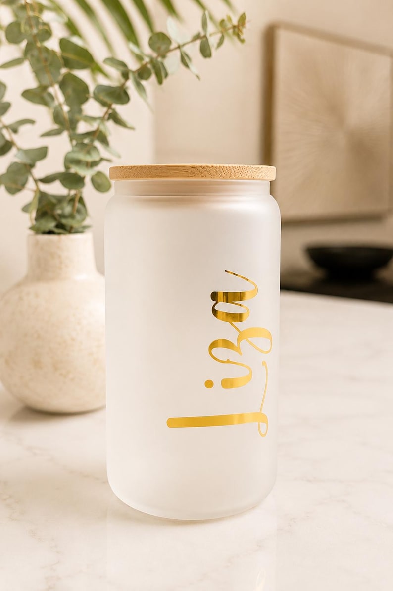 May include: A cylindrical frosted glass tumbler with a light-colored bamboo lid. The glass features the name Liza written in a metallic gold cursive script font on the front surface. The tumbler has a smooth, matte finish and a straight-sided design.