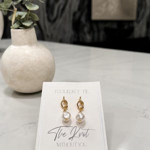 May include: A pair of gold-toned knot earrings with white pearl-like drops attached to a white display card. The card features the printed text I COULDN'T TIE The Knot WITHOUT YOU in black ink, centered above and below the earrings.