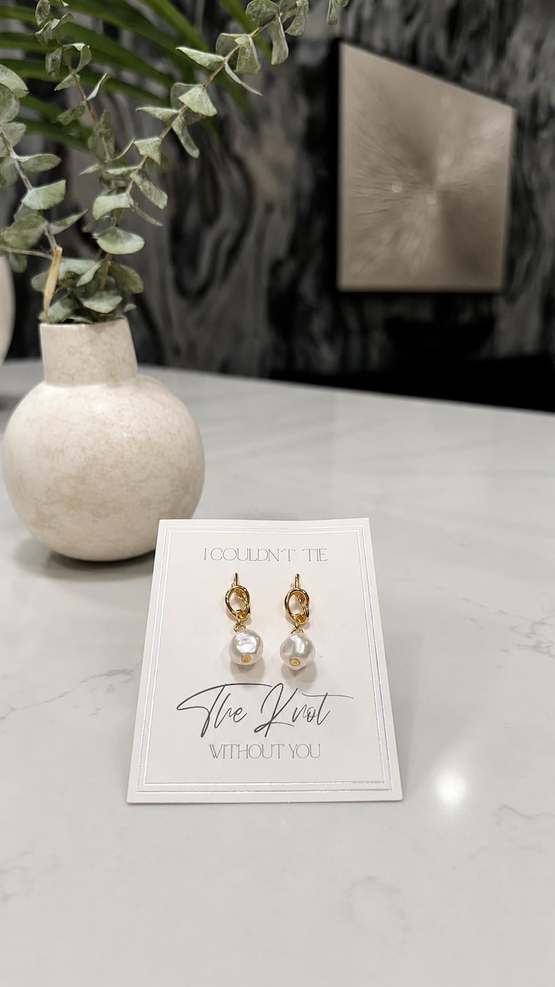 May include: A pair of gold-toned knot earrings with white pearl-like drops attached to a white display card. The card features the printed text I COULDN'T TIE The Knot WITHOUT YOU in black ink, centered above and below the earrings.