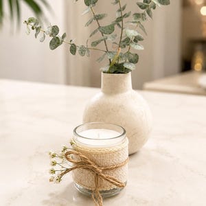 May include: A clear glass candle jar wrapped in a beige burlap ribbon with a jute twine bow and small white flowers attached to the front. Behind the candle sits a round, off-white ceramic vase containing several stems of green eucalyptus.