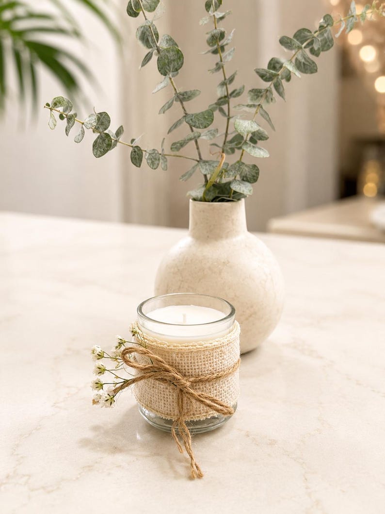May include: A clear glass candle jar wrapped in a beige burlap ribbon with a jute twine bow and small white flowers attached to the front. Behind the candle sits a round, off-white ceramic vase containing several stems of green eucalyptus.