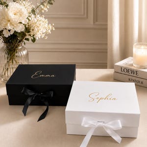 May include: Two rectangular gift boxes with lids, one matte black with the name Emma in gold script and a black ribbon, and one matte white with the name Sophia in gold script and a white ribbon, both featuring a magnetic closure mechanism.