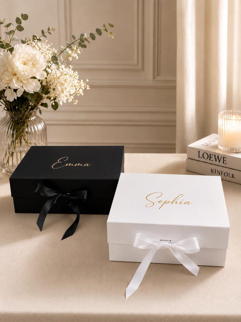 May include: Two rectangular gift boxes with lids, one matte black with the name Emma in gold script and a black ribbon, and one matte white with the name Sophia in gold script and a white ribbon, both featuring a magnetic closure mechanism.