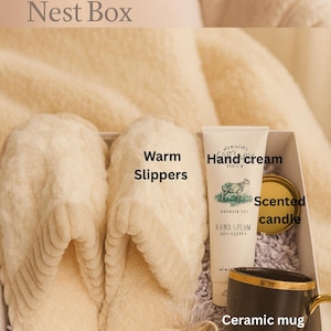 May include: A gift box labeled "Calm Nest Box" containing cream-colored warm slippers, hand cream, a scented candle, a ceramic mug, and a small bag of treats. The mug has a gold handle and trim. The box is filled with shredded paper.