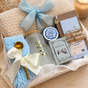 May include: A gift set containing a beige knit blanket with a light blue ribbon, a pair of light blue cable-knit socks with a gold spoon, a frosted glass tumbler with gold text, a small candle, a notebook, and a box of Moroccan Mint tea.