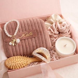May include: A pink gift box filled with self-care items. Includes a pearl bracelet, a small pink pouch with "Kate" embroidered on it, a wooden hairbrush, a candle, and satin scrunchies. The box is lined with pink paper and ribbon.