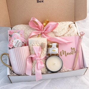 May include: A gift box labeled "Calm Nest Box" filled with self-care items. Includes a pink mug with a gold handle, a journal with "Mom" written on it, a pen, a candle, honey, tea, and a knitted cozy. All items are tied with pink ribbons.