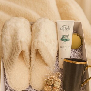 May include: A gift box containing cream-colored slippers, a tube of goat's milk hand cream, a black mug with a gold handle, and a small bag of gold-wrapped chocolates. The box is lined with shredded paper.