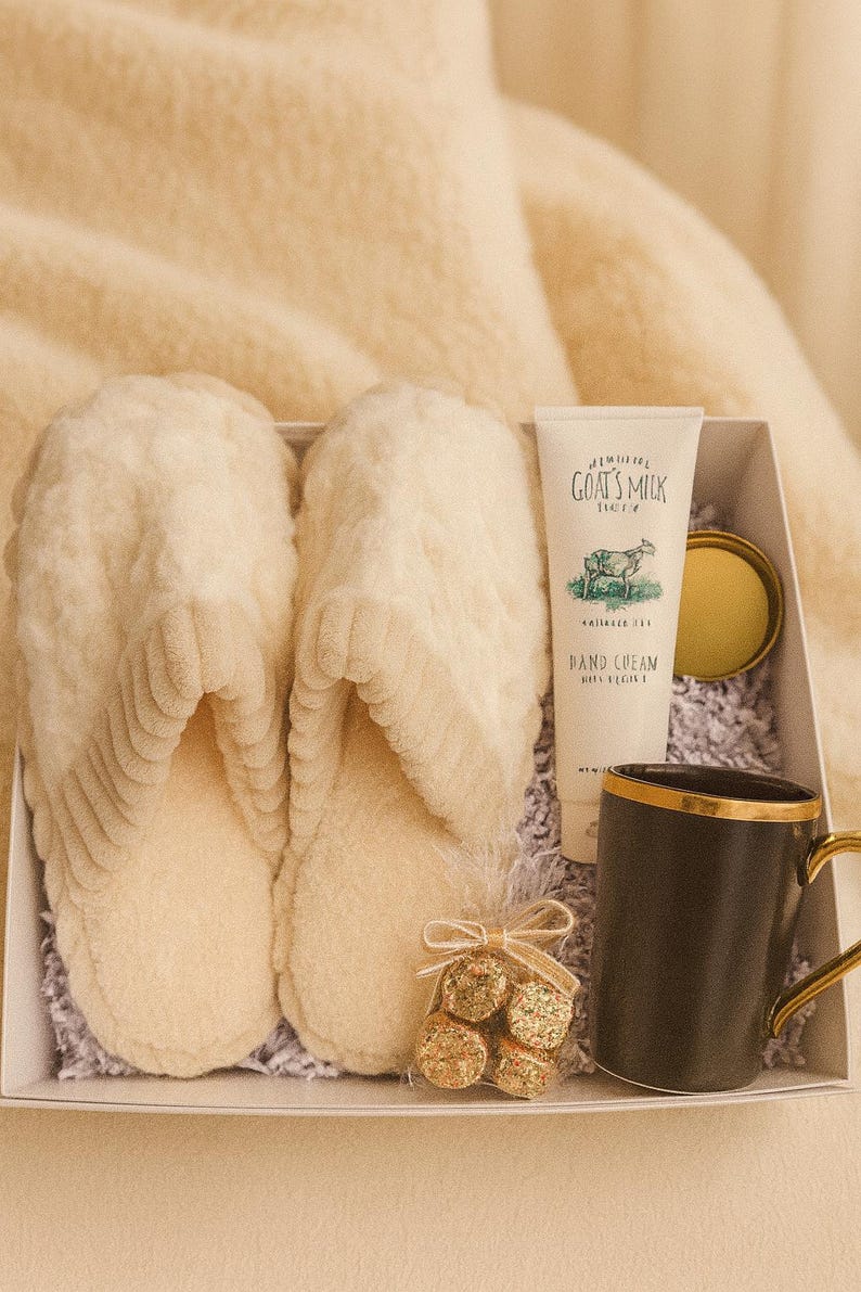 May include: A gift box containing cream-colored slippers, a tube of goat's milk hand cream, a black mug with a gold handle, and a small bag of gold-wrapped chocolates. The box is lined with shredded paper.