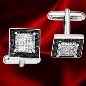 Puede incluir: Un par de gemelos plateados con diseño cuadrado. Cada gemelo presenta un cuadrado central de pequeñas piedras transparentes, rodeado por un borde de piedras negras. Sobre un fondo rojo oscuro.