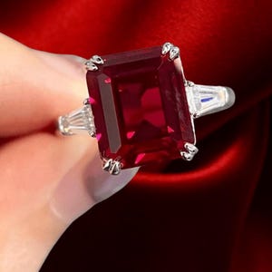 Puede incluir: Un anillo plateado con una gran gema rectangular de color rojo intenso como pieza central. Dos gemas blancas rectangulares más pequeñas flanquean la piedra principal. El anillo se encuentra sobre un fondo rojo intenso.