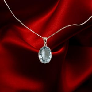 K&ouml;nnte beinhalten: Eine silberne Halskette mit einem hellblauen, ovalen Anh&auml;nger. Der Anh&auml;nger h&auml;ngt an einer feinen Kette und ist vor einem roten Satinstoff platziert. Der Edelstein hat eine subtile, durchscheinende Qualit&auml;t.
