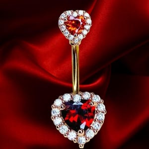 May include: A gold-coloured belly button ring featuring two heart-shaped gemstones. The top heart is orange, and the bottom heart is red. Both hearts are encircled by a halo of small, clear gemstones. The background is a deep red, silky fabric.
