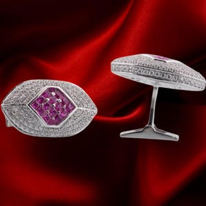 May include: Two silver cufflinks with a rugby ball shape. Each cufflink has a central square of small, pink gemstones. The edges are encrusted with small, clear gemstones. The cufflinks are displayed on a red satin background.