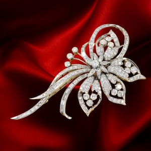 May include: A diamond and gold floral brooch. The brooch features a central flower design with multiple diamond accents and a curved stem. The brooch is set against a red background.