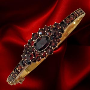Könnte beinhalten: Ein goldfarbenes Armband mit Scharnier, das einen zentralen ovalen Granatstein umgeben von kleineren Granaten aufweist. Das Armband hat ein sich wiederholendes Muster aus kleinen Granaten an den Rändern, vor rotem Hintergrund.
