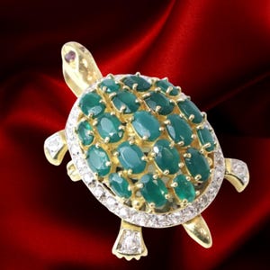 May include: A gold-toned turtle brooch with a shell of oval-cut emeralds. The brooch features diamond accents around the shell and on the feet. A small ruby is set on the turtle's head. The background is a deep red.