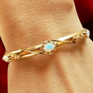 May include: A gold bangle bracelet with an oval opal stone in the center, surrounded by a floral design. The bracelet has additional opal stones at each end and is adorned with delicate gold detailing.
