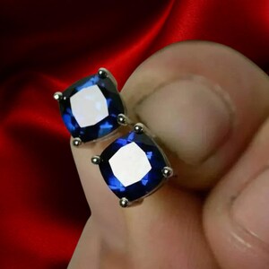 May include: A pair of square-cut, deep blue gemstone stud earrings. Each earring features a silver-colored setting with four small prongs holding the gemstone in place. The gemstones have a bright, reflective surface.