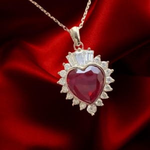 May include: A silver necklace with a heart-shaped, deep red gemstone pendant. The pendant is surrounded by smaller, clear gemstones and has a crown-like design at the top. The necklace hangs against a rich, red fabric background.