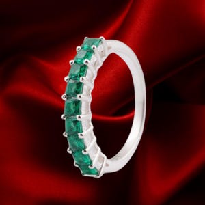 May include: A silver ring with a line of emerald-cut green gemstones. The ring's band is silver, and the gemstones are set closely together. The background is a deep red, creating a contrast with the ring's colors.