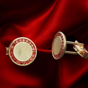 Puede incluir: Gemelos dorados con diseño circular. Cada gemelo presenta un círculo central texturizado rodeado de una hilera de piedras rojas. Sobre un fondo rojo intenso.
